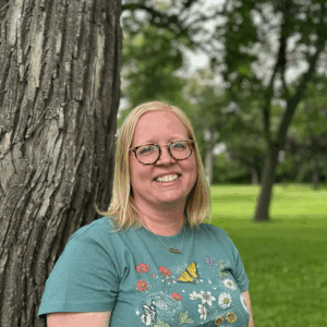 Lyndsay smiles and stands in front of a tree wearing a blue shirt.